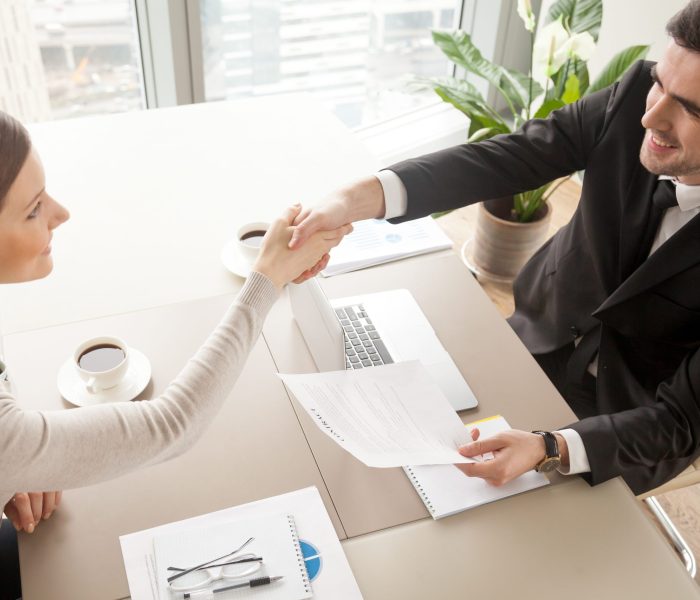 Smiling female and male business leaders handshaking over desk after or before signing contract in office. Happy businesswoman congratulating businessman with concluding profitable agreement. Top view